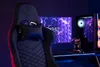 A gaming chair is positioned in front of a computer setup with a monitor displaying a hexagonal pattern and a computer tower with RGB lighting. Produktbild-Vorschau