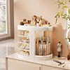 A white rotating makeup organizer filled with various cosmetic products sits on a dresser. Náhled obrázku produktu