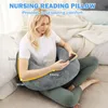 A woman is sitting on a bed, using a U-shaped pillow for support while looking at her phone. The pillow is gray and wraps around her. Produktbild-Vorschau