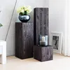 A collection of wooden pedestals of varying heights, decorated with a vase of flowers and a candle lantern, in a room with a concrete wall and white curtains. Anteprima dell’immagine prodotto