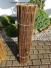 A rolled-up willow mat is standing upright on a paved patio, with green bushes and gravel in the background. Produktbild-Vorschau