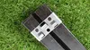A close-up shot of a black composite material plank with a silver metal clip attached, lying on a green grassy surface. Produktbild-Vorschau