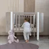 A baby standing next to a crib with a teddy bear in the foreground. Produktbild-Vorschau