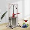A stainless steel apple press with a red hydraulic jack, set against a light background with a plant on the left and a glass of red wine and grapes on the right. Náhled obrázku produktu