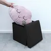 A hand is placing a pink cloud-shaped pillow with closed eyes and a small grey stuffed hippo into an open black storage ottoman. Náhľad obrázku produktu