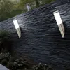 Two modern outdoor wall lights mounted on a dark stone wall, with lush green plants in the foreground. Produktbild-Vorschau