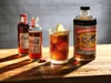 A cocktail and two bottles of Coca-Cola are arranged on a wooden surface, with a bottle of Shanky's Whip Irish Whisky Liqueur in the background. Produktbild-Vorschau