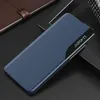 A blue flip case for a smartphone is displayed on a dark surface, with a portion of a brown notebook visible in the upper left corner. Produktbild-Vorschau