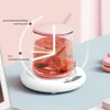 A pink-lidded glass mug filled with a pink beverage and ice is placed on a white electric mug warmer. The scene is set on a light blue surface with a pink macaron in the background. Produktbild-Vorschau