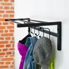 A black metal coat rack mounted on a white wall, with several items of clothing and a hat hanging on it. A brick wall is visible to the left. Produktbild-Vorschau