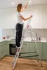 A woman is standing on a ladder in a kitchen, changing a lightbulb. Anteprima dell’immagine prodotto