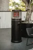 A black patio heater stands on a tiled patio next to a large potted tree and a chair. Produktbild-Vorschau