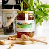 A jar of honey and almonds on a wooden board, with a plant in the background. Produktbild-Vorschau