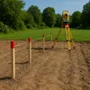 Surveying equipment and wooden stakes are set up on a dirt field, with a grassy area and trees in the background. Produktbild-Vorschau