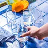 A hand reaches for a glass of water on a blue tiled surface, with oranges and a bottle in the background. Voir les images du produit