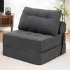 A dark gray foldable sofa bed is shown in a room setting, with a wooden cabinet and a plant in the background. Náhled obrázku produktu