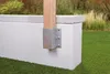 A close-up of a wooden post secured to a textured white concrete structure with a metal bracket and bolts. The background shows green grass and some dry ornamental grasses. Voir les images du produit