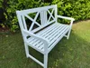 A white wooden garden bench sits on a grassy lawn in front of a green hedge. Produktbild-Vorschau