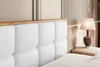 A close-up view of a white upholstered headboard with a wooden trim, next to a bedside table with a lamp. Produktbild-Vorschau