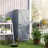 A grey wooden garden cabinet with double doors stands on a green lawn next to a white railing and a white wooden wall. Several potted plants are visible on a white shelf to the left, and more plants are on the railing and in front of the cabinet. Náhľad obrázku produktu