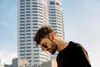 A man wearing sunglasses and earbuds stands in front of a modern building. Produktbild-Vorschau