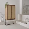 A tall, narrow cabinet with two doors made of wood and rattan, standing on a black metal frame. It is placed in a minimalist, brightly lit room with white walls and tiered platforms. Produktbild-Vorschau