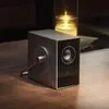 A speaker is placed on a wooden surface next to a lit candle in a glass holder. Produktbild-Vorschau