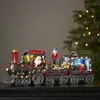 A festive Christmas train decoration with Santa, a nutcracker, and gifts, illuminated by LED lights, set against a blurred background with Christmas tree lights. Podgląd zdjęcia produktu