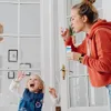 Frau in orangefarbenem Kapuzenpullover bläst Blasen aus einer kleinen blauen Flasche, ein glückliches Kind greift nach ihnen Náhled obrázku produktu