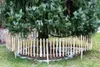 A small wooden fence surrounds the base of a large evergreen tree in a grassy yard. Produktbild-Vorschau