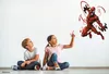 Two children sitting on the floor looking up at a wall sticker of a superhero. Voir les images du produit