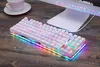 A white gaming keyboard with RGB lighting is placed on a wooden desk next to a white cup and saucer. A computer monitor is visible in the background. Produktbild-Vorschau