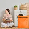 A woman is sitting on the floor next to a washing machine, holding a stack of folded clothes. An orange laundry bag with a zipper is placed in front of the washing machine. Produktbild-Vorschau