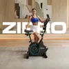 A woman is posing next to a stationary bike in a modern living room setting. Produktbild-Vorschau
