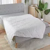 A white quilted blanket is laid out on a bed with a grey headboard and grey fitted sheet. Produktbild-Vorschau