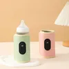 Two portable bottle warmers, one green and one pink, are displayed on a white tray against a light beige background. A white pleated lampshade is visible in the upper right corner. Produktbild-Vorschau