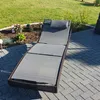A grey chaise lounge with a black cushion is placed on a paved patio next to a garden bed with plants and grass. Produktbild-Vorschau
