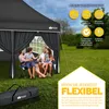 A family is sitting under a black pop-up gazebo in a grassy outdoor setting. Produktbild-Vorschau