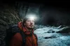 A man wearing a headlamp and backpack stands by a stream in a dark forest. Podgląd zdjęcia produktu