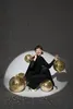A woman in a black sequined outfit is crouching surrounded by several gold disco balls, with a spotlight illuminating the scene and creating a speckled pattern on the background. Anteprima dell’immagine prodotto