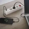 A white power strip is plugged into a wall outlet and connected to a laptop and a smartphone, both of which are charging. The smartphone screen displays a charging icon. Produktbild-Vorschau