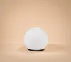 A white spherical lamp with a black base sits on a peach-colored surface. Produktbild-Vorschau