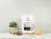 A white coffee maker with a glass carafe filled with coffee, a yellow mug, a small plant, and a jar of coffee grounds on a marble countertop. Produktbild-Vorschau
