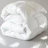 A folded white duvet or comforter is shown on a light gray background with several white feathers scattered around it. Produktbild-Vorschau