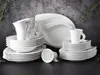 A collection of white porcelain dinnerware, including plates, bowls, and cups, arranged on a dark surface against a textured gray background with green foliage accents. Produktbild-Vorschau