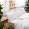 A white bed with multiple pillows and a duvet, with a wooden bedside table and plants in the foreground. Anteprima dell’immagine prodotto