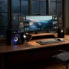 A computer desk setup with a large monitor, speakers, keyboard, and mouse, set against a window with a view of mountains at dusk. Produktbild-Vorschau