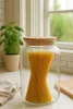 A glass storage jar filled with spaghetti, placed on a kitchen counter next to a potted plant and a dishtowel. Produktbild-Vorschau