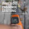 A hand holding an orange and black leaf blower with a digital display showing power levels and icons. The background is blurred and shows an outdoor setting with some leaves. Podgląd zdjęcia produktu