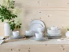 A collection of white porcelain dinnerware with gold trim, including plates, bowls, and cups, arranged on a wooden surface with a vase of white flowers. Náhľad obrázku produktu